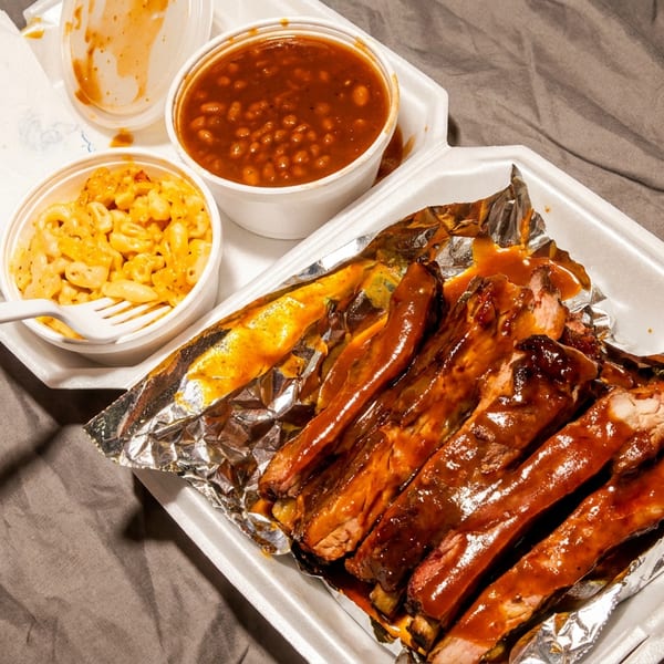 Saucy BBQ ribs, mac and cheese, and baked beans in a takeout container.